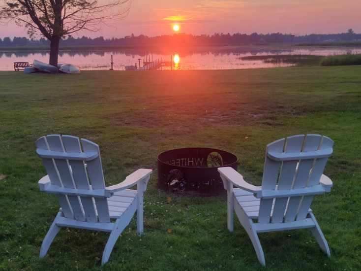 Two white Adirondack chairs and a fire pit facing the sunset over the bay.